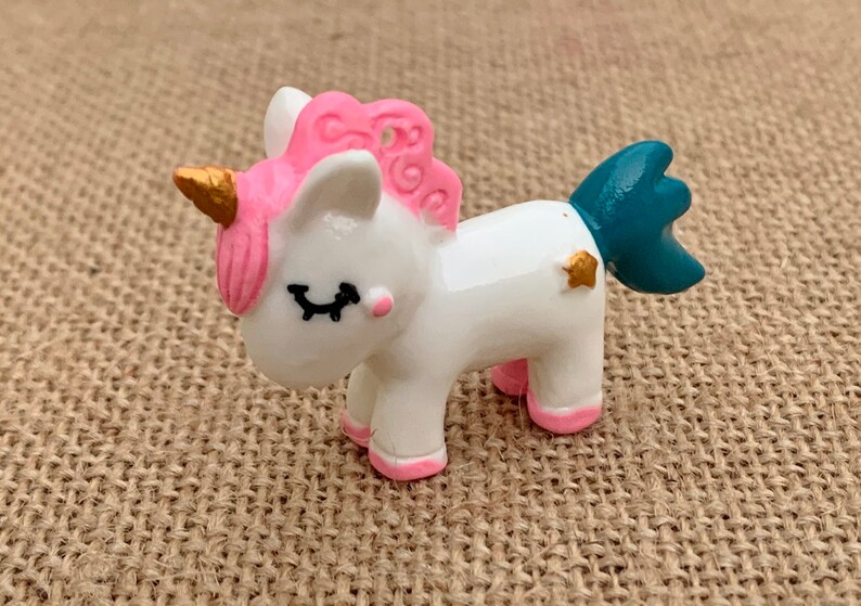 May include: A small white unicorn figurine with pink hair, a gold horn, and a teal tail. The unicorn has closed eyes and a gold star on its side.