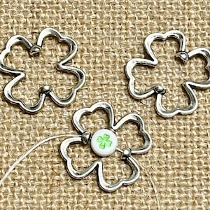 May include: Three silver metal four-leaf clover charms, one with a green shamrock design in the center. The charms are on a burlap background.