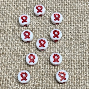 May include: Ten white round beads with a red ribbon design. The beads are arranged in a circle on a brown burlap background.