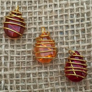 May include: Three gemstone pendants with gold wire wrapping. The pendants are teardrop-shaped and vary in color: red, orange, and a darker red. The pendants are displayed on a textured, tan fabric background.