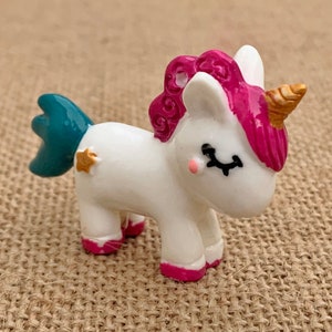 May include: A small white unicorn figurine with a pink mane and tail, a teal blue tail, and a gold horn. The unicorn has closed eyes and a gold star on its side.
