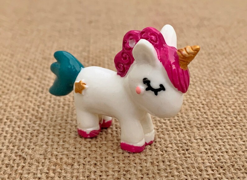 May include: A small white unicorn figurine with a pink mane and tail, a teal blue tail, and a gold horn. The unicorn has closed eyes and a gold star on its side.
