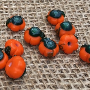 May include: A collection of orange and green glass pumpkin beads. The beads are about 1 inch in diameter and have a glossy finish.