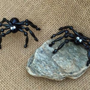 May include: Two black beaded spiders with googly eyes. The spiders are crafted with black beads and have a shiny finish. One spider is on a grey rock and the other is on a burlap background.