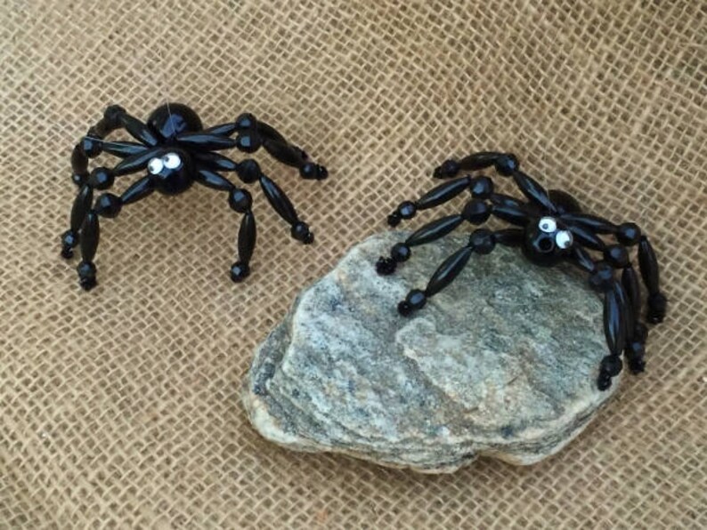 May include: Two black beaded spiders with googly eyes. The spiders are crafted with black beads and have a shiny finish. One spider is on a grey rock and the other is on a burlap background.