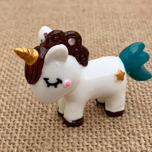 May include: A small white unicorn figurine with a brown mane and tail, a gold horn, and a teal tail. The unicorn has a gold star on its side and closed eyes with pink accents.