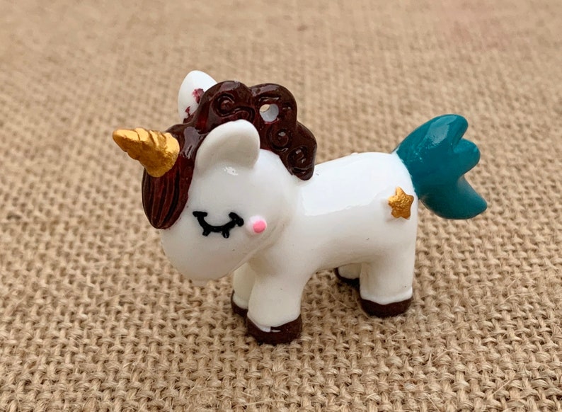 May include: A small white unicorn figurine with a brown mane and tail, a gold horn, and a teal tail. The unicorn has a gold star on its side and closed eyes with pink accents.