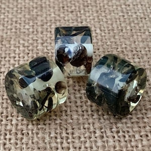 May include: Three clear, square-shaped beads with dark, mottled inclusions. The beads are arranged on a textured, tan surface. The inclusions appear to be dark brown and black, creating a unique pattern within each bead. The beads are translucent.