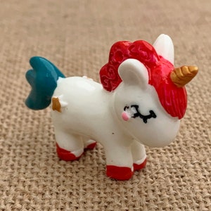 May include: A small white unicorn figurine with a red mane and tail, blue hooves, and a gold horn. The unicorn is standing on a brown hessian surface.