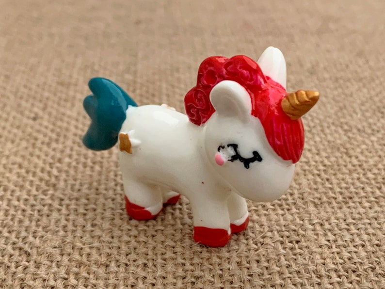 May include: A small white unicorn figurine with a red mane and tail, blue hooves, and a gold horn. The unicorn is standing on a brown hessian surface.