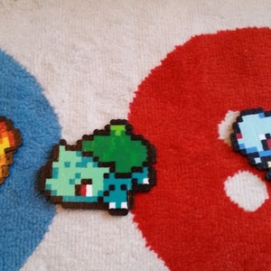 May include: Three pixel art figures of Pokemon characters: Charmander, Bulbasaur, and Squirtle. Charmander is orange and red with a flame tail. Bulbasaur is green with a brown shell and a green plant on its back. Squirtle is blue with a brown shell and a blue tail.