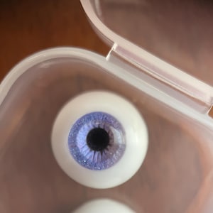 May include: A pair of realistic, white-based prosthetic eyes with a detailed, light blue iris and a black pupil. The eyes are in a clear plastic container, suggesting they are new or unused. The iris has a textured appearance.
