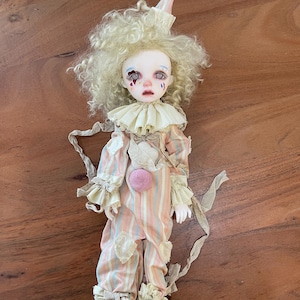 May include: A handmade clown doll with a pale face, curly blonde hair, and a pointed hat. The doll wears a striped pink and blue jumpsuit with ruffled sleeves and a large collar. A pink pom-pom adorns the doll's chest.
