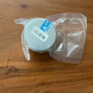 May include: A small, round, silver-colored container with the text "KYAN'S WORKS 奶油" on a white label. The container is sealed in a clear plastic package. The background is a wooden surface.