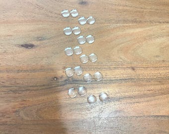 May include: Clear, round glass gems arranged on a wooden surface. The gems are small and appear to be smooth, with a slight dome shape. The wooden surface has a natural wood grain pattern.