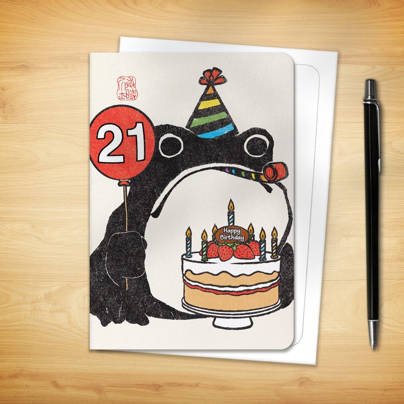 May include: Birthday card featuring a black cartoon frog holding a red balloon with the number 21. The frog wears a party hat and holds a cake with strawberries and candles. The words "Happy Birthday" are on the cake. A black pen and white envelope are next to the card.