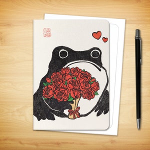 May include: Greeting card featuring a black frog holding a bouquet of red roses, with two red hearts above. The card has a cream background and a red stamp in the upper left corner. A white envelope and a black pen are next to the card.
