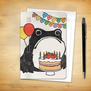 May include: A birthday card featuring a black frog holding balloons and a cake with strawberries. The card has a banner that says "Happy Birthday". A white envelope and a black pen are next to the card on a wooden surface.