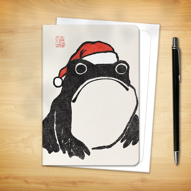 May include: Greeting card featuring a black and white illustration of a frog wearing a red Santa hat. The card has a rounded edge and is accompanied by a white envelope and a black pen, all set on a wooden surface.