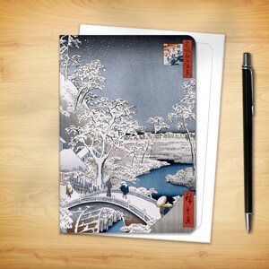 May include: Greeting card featuring a Japanese woodblock print of a snowy landscape with a bridge, river, and figures. The print is in shades of blue, white, and gray, with Japanese text in red. A black pen rests on a wooden surface.
