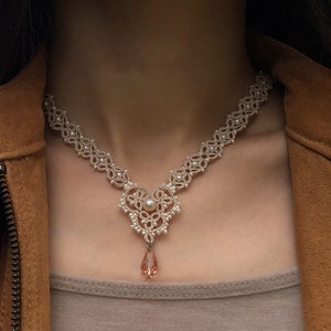 May include: A delicate white beaded necklace with a pearlescent pendant and a pink crystal drop. The necklace is worn by a person with brown hair and a brown leather jacket.