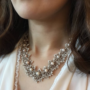 以下が含まれることがあります： パールがあしらわれた白いクロシェネックレス。ネックレスは繊細で複雑なレースワークで作られており、花柄のデザインが特徴です。