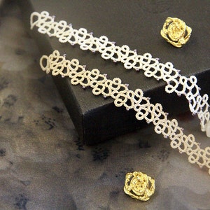 Może przedstawiać: Dwie kremowe bransoletki o otwartym splocie z delikatnym, kwiatowym wzorem, zaakcentowane małymi fioletowymi koralikami. Bransoletki są prezentowane na ciemnej powierzchni z trzema złotymi broszkami w kształcie róży.