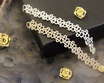 May include: Two cream-coloured, open-weave bracelets with a delicate, floral-like pattern, accented with small purple beads. The bracelets are displayed on a dark surface with three gold-coloured rose-shaped brooches.