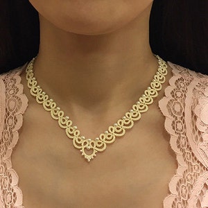 Könnte beinhalten: Eine zarte, goldfarbene Halskette mit einem aufwendigen, durchbrochenen Design. Die Halskette weist ein sich wiederholendes Muster aus verbundenen Schlaufen und einen zentralen Anhänger auf. Die Halskette wird über einem rosa Spitzenkleid getragen.