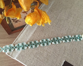 May include: A handmade bracelet with a teal and white floral lace design. The bracelet is decorated with small white beads and has a button closure. The bracelet is approximately 18 cm long.