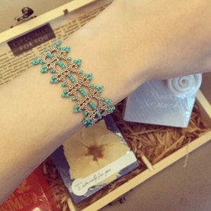 May include: A delicate beige and turquoise beaded bracelet with a lacy design. The bracelet is displayed on a person's wrist.