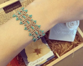 May include: A delicate beige and turquoise beaded bracelet with a lacy design. The bracelet is displayed on a person's wrist.