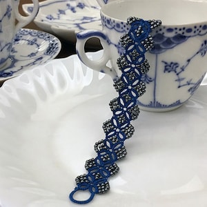 Pode incluir: Uma pulseira azul escura com contas e um design floral, colocada num prato branco. A pulseira é feita de fio azul escuro e pequenas contas prateadas. No fundo, xícaras e pires brancos com padrões florais azuis.
