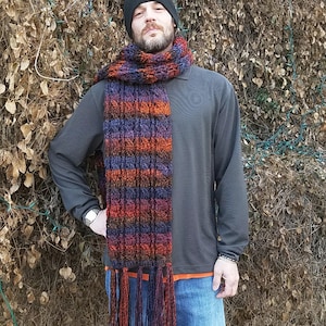 May include: A handmade, fringed scarf in shades of orange, purple, and blue. The scarf is worn with a black beanie featuring a red maple leaf, a gray long-sleeve shirt, and blue jeans. The scarf is a cozy accessory.