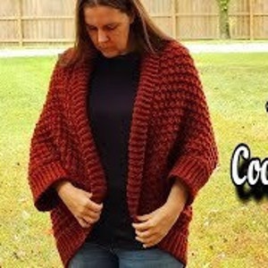 May include: A red crocheted cardigan with a cocoon shape and a textured stitch pattern. The cardigan is open in the front and has long sleeves.