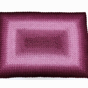 May include: A rectangular crocheted rug with a gradient of pink and burgundy colors. The rug has a square design in the center.