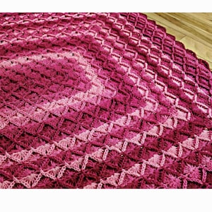 Puede incluir: Una manta de ganchillo con un patrón de degradado rosa y rojo. La manta está hecha con un punto de diamante y tiene un borde festoneado.