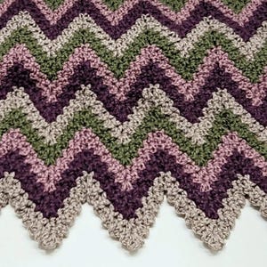 Peut inclure: Une couverture en crochet chevron dans des tons de violet, rose et vert. La couverture a un bord festonné.