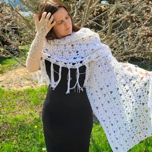 May include: A white crochet shawl with a heart pattern and fringe. The shawl is draped over a person wearing a black dress.