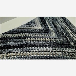May include: A close-up of a crocheted shawl with a gradient of gray and black yarn. The shawl has a textured pattern with a repeating motif.