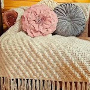 以下が含まれることがあります： フリンジ付きのクリーム色のクロシェ編み毛布が、豪華なベルベットの肘掛け椅子にかけられています。ピンクとグレーの2つの装飾的なクッションが毛布の上に置かれています。