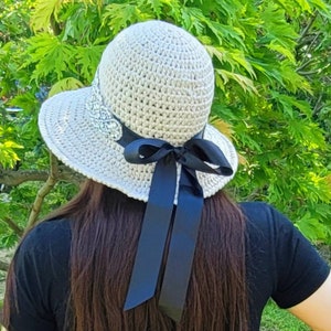 May include: A white crocheted sun hat with a black ribbon tied in a bow at the back. The hat has a decorative embellishment on the front.