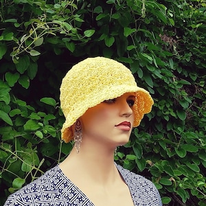 Puede incluir: Un sombrero de sol amarillo tejido a ganchillo con ala, exhibido en un maniquí. El sombrero tiene un diseño texturizado y de tejido abierto. El maniquí lleva una blusa estampada en azul y blanco, sobre un fondo de follaje verde.