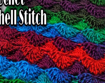 May include: A close-up of a colourful crocheted fabric with a shell stitch pattern. The fabric is made with different shades of green, blue, red, and purple yarn.