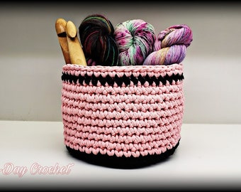 Puede incluir: Cesta de crochet rosa con ribete negro, llena de hilo y agujas de tejer.  "Bag-O-Day Crochet" está escrito debajo de la cesta.