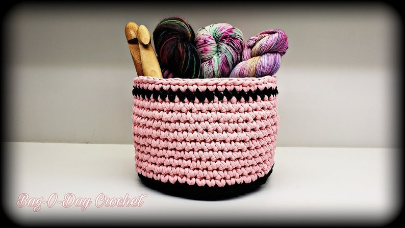 May include: Pink crocheted basket with black trim, filled with yarn and knitting needles.  "Bag-O-Day Crochet" is written below the basket.
