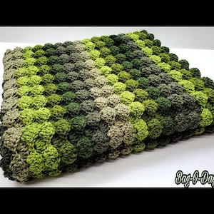 Puede incluir: Una manta de crochet verde, marrón y beige con una superficie texturizada y abultada. La manta está hecha con un patrón repetitivo de pequeños puntos redondos.