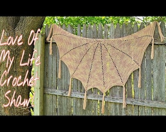May include: A crocheted shawl with a scalloped edge and fringe. The shawl is a light beige color with a subtle pink and yellow ombre effect. The shawl is draped over a wooden fence.