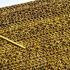 May include: Close-up of a gold crochet fabric with a repeating pattern of small flower shapes. A gold crochet hook is lying on top of the fabric.
