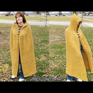 Op de afbeelding: Een gele gehaakte kap met bruine knopen. De cape is lang en vloeiend, en de capuchon is groot en ruim. De cape wordt gedragen door een persoon die in een grasveld staat.